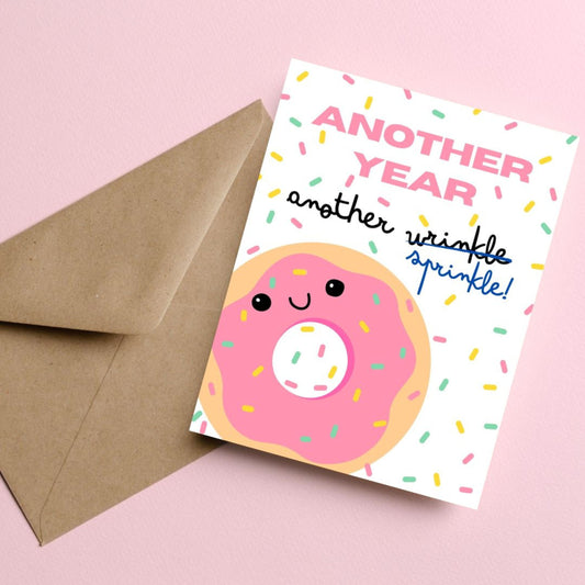 Birthday card with a donut covered in sprinkles on a sprinkle background, featuring the funny phrase "Another Year, Another Sprinkles" with "sprinkles" crossed out and replaced by "wrinkles." Perfect for celebrating aging with humor and sweetness.