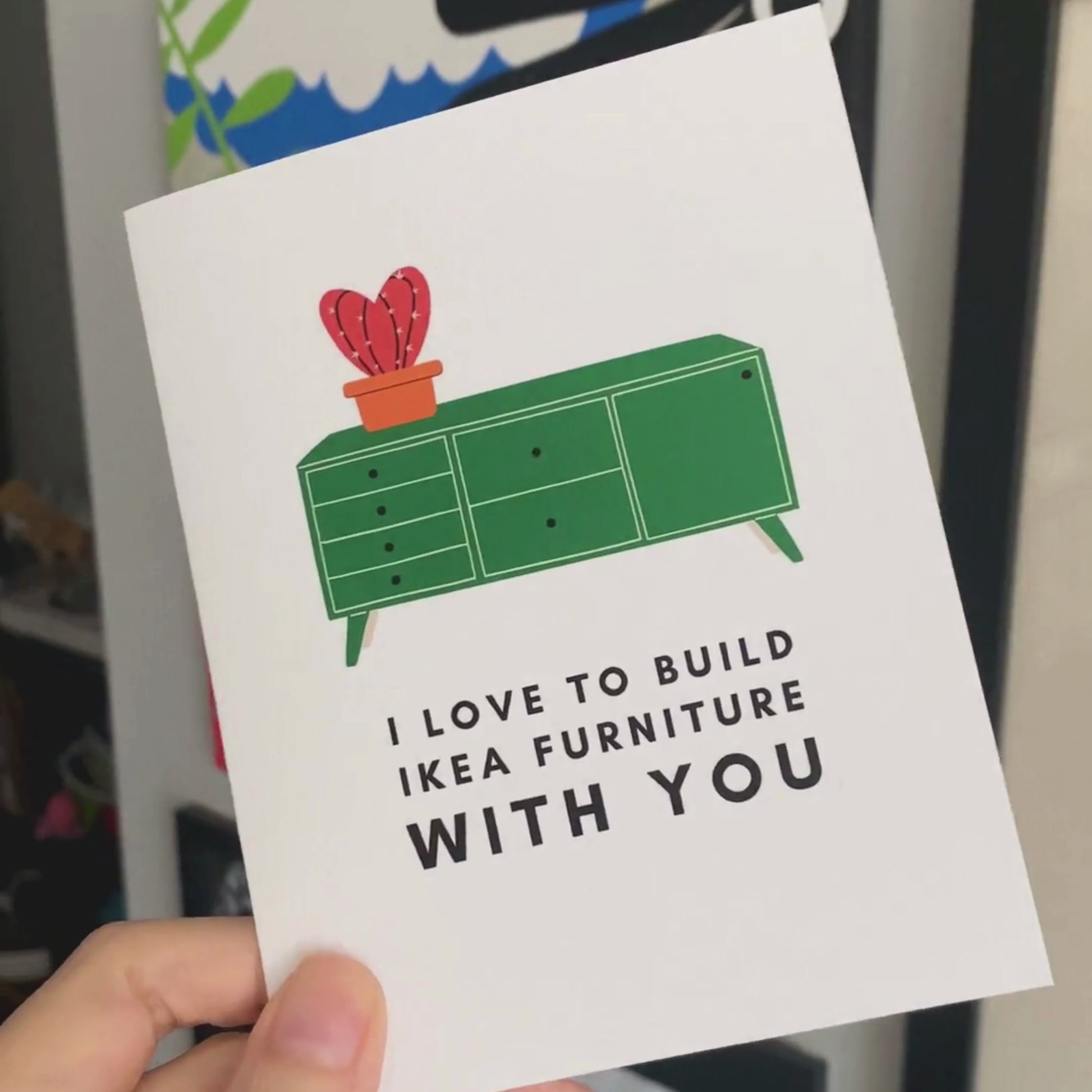 Greeting card with the phrase I Love to Build IKEA Furniture With You featuring a drawing of a green TV stand with a vase holding a red cactus on top. Perfect for celebrating relationships with humor and a nod to DIY adventures.
