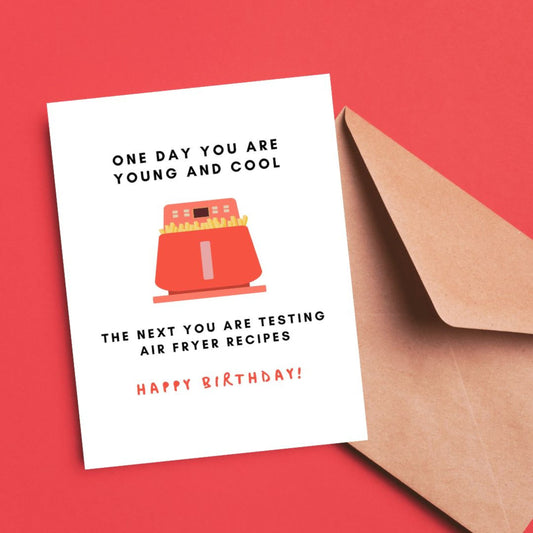 Birthday greeting card with the phrase One Day You Are Young And Cool, The Next You Are Testing Air Fryer Recipes featuring a drawing of a red air fryer and Happy Birthday written at the bottom. Perfect for celebrating with humor and relatable twist.