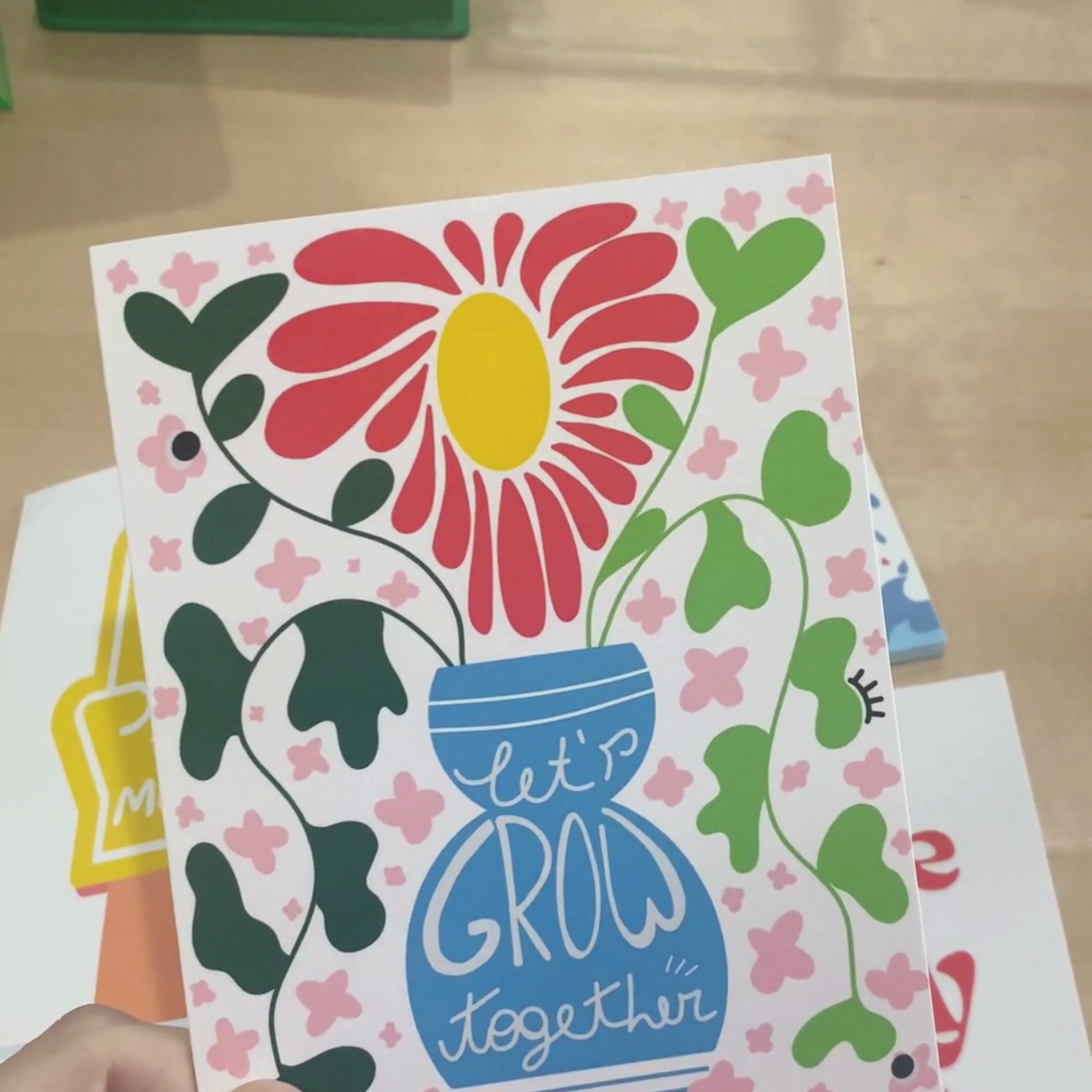 Greeting card with original drawing of blue vase with greenery and big yellow and red flower, phrase Let's Grow Together inside the vase.