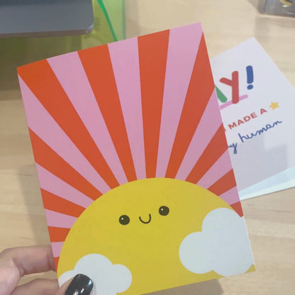  Sunshine Greeting Card with a drawing of a big yellow sun with a smiling face, pink and red rays, and two clouds. Blank inside, perfect for spreading cheerful vibes and warm wishes.